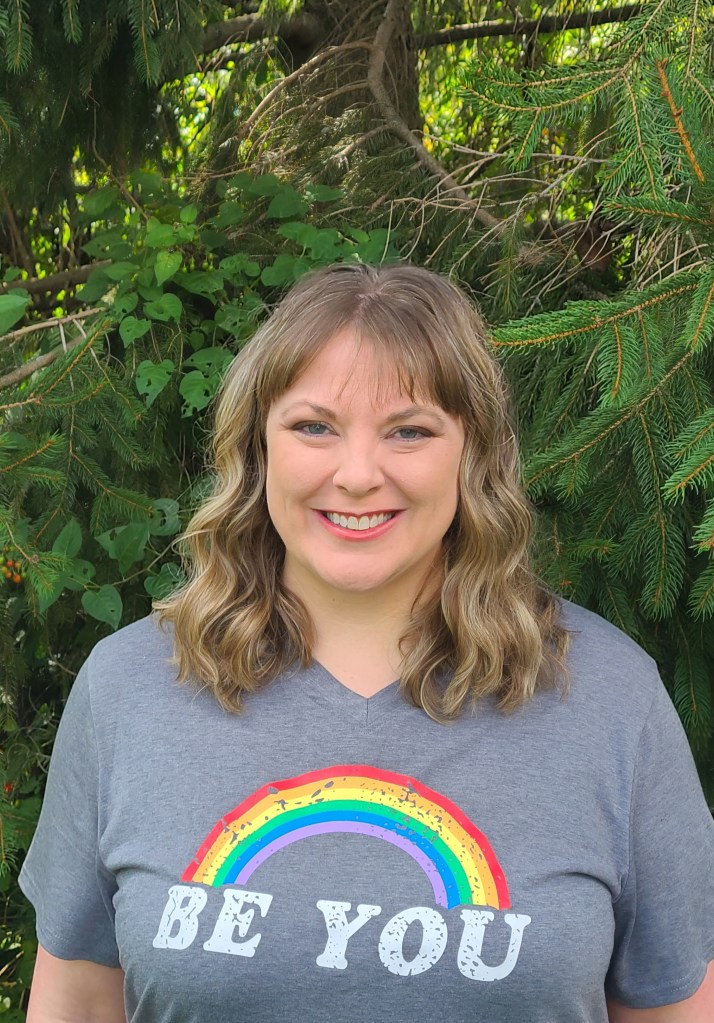 Image description: The photo depicts an adult female, Katie Wine-Appleton. She is wearing a T-shirt featuring a rainbow graphic with the words 'Be you'.